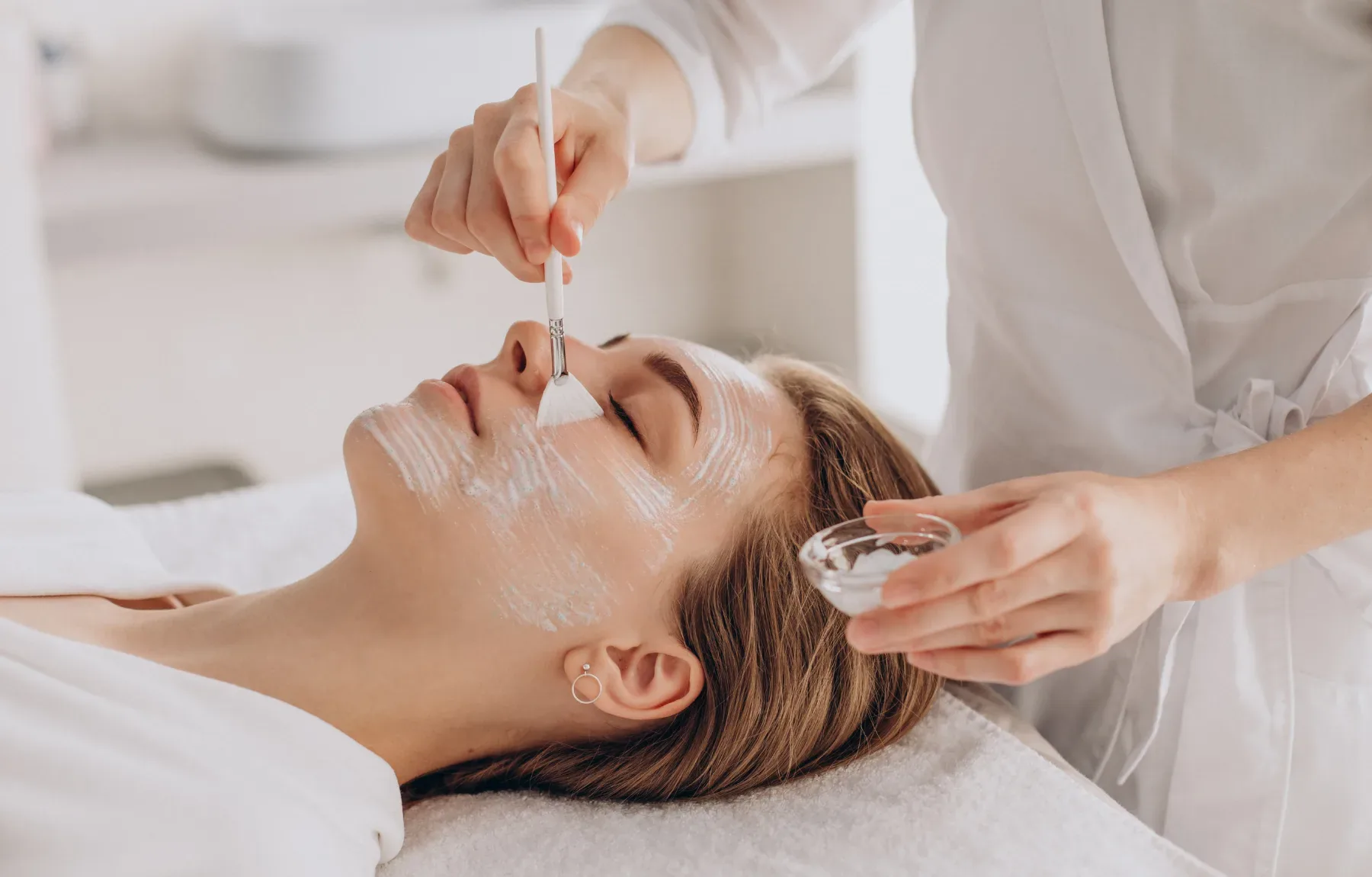 A woman lies with her eyes closed while a provider brushes a white mask or peel product onto her face with a small applicator