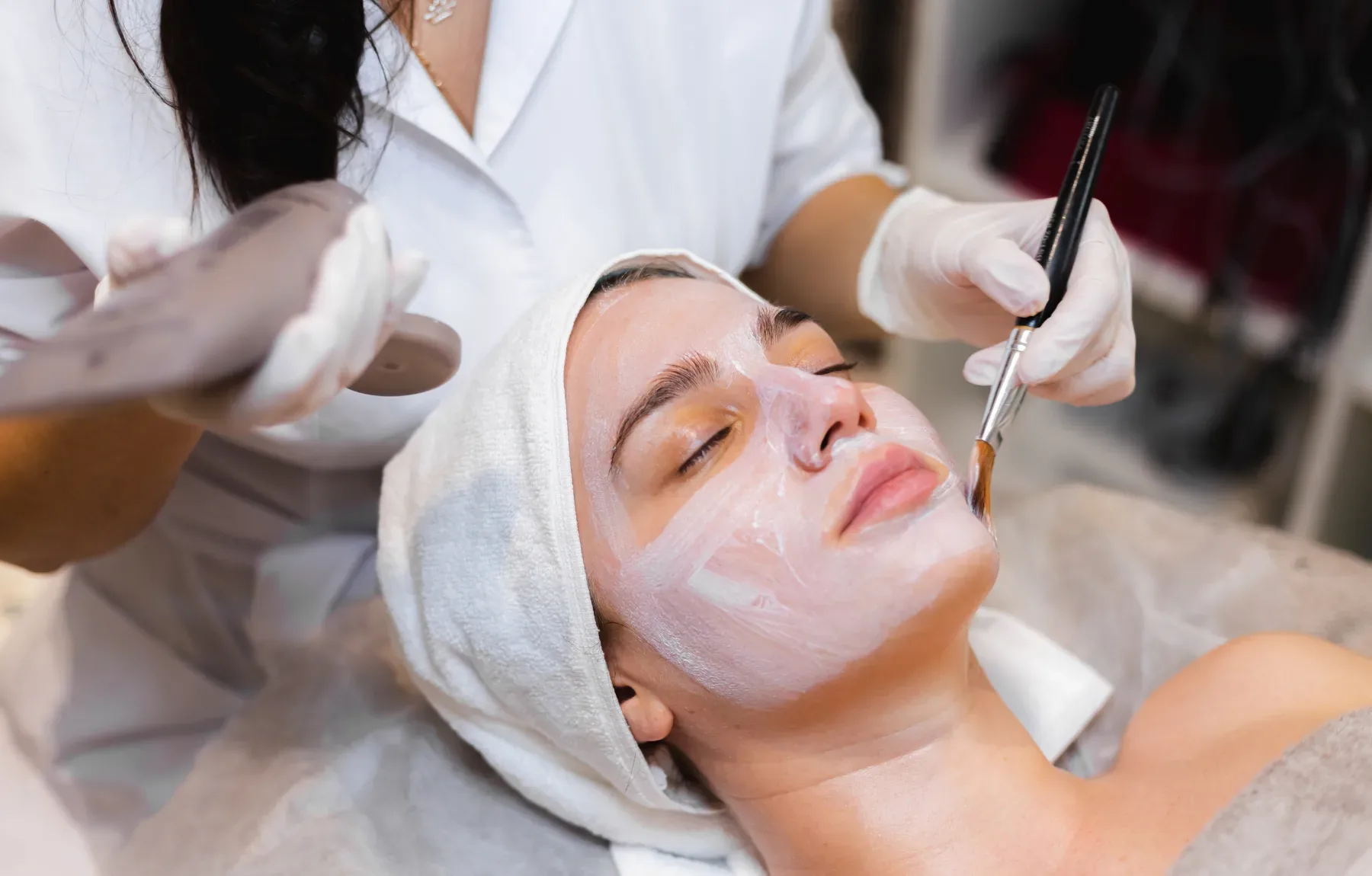 A woman with a white towel wrapped around her head lies with her eyes closed while a gloved hand brushes a peach-toned mask o